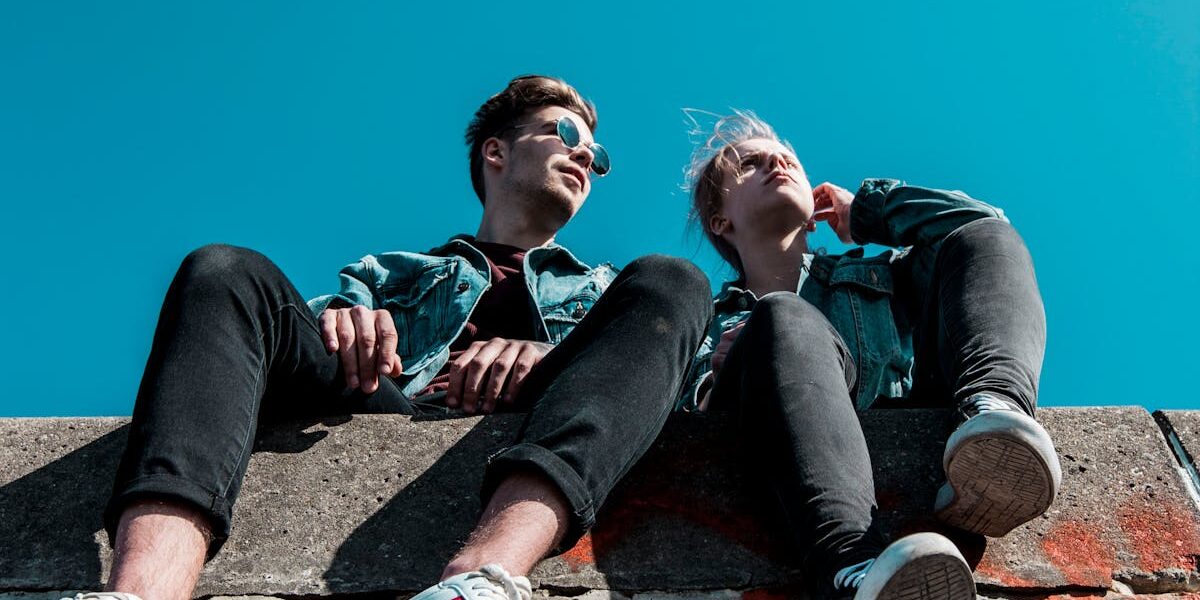Two young people sitting on a stone wall under a vibrant blue sky, enjoying a sunny day together.
