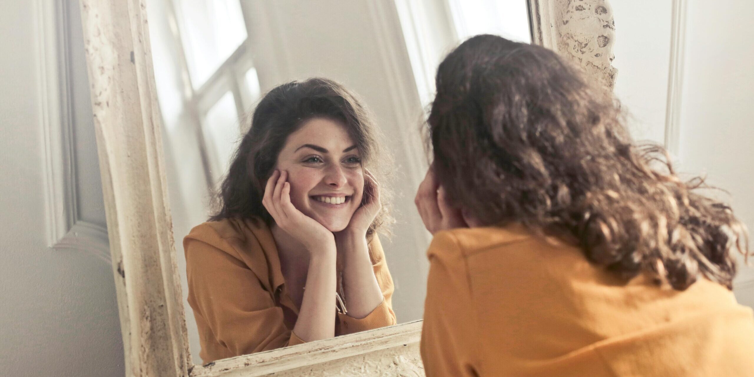 A cheerful woman smiles at her reflection in a vintage-style mirror, exuding positivity and warmth.